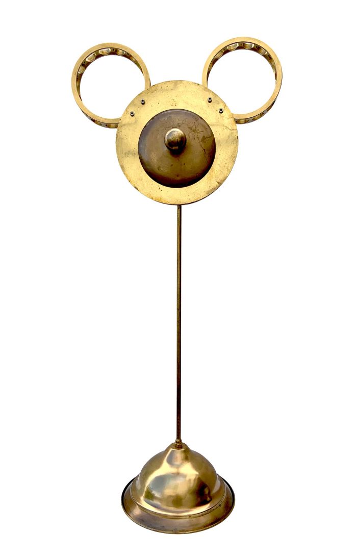escultura amb la cara formada per una rodona amb un pom per mas i uns cercels industrials per orelles, te una base d'on surf una tija que soporta el cap
