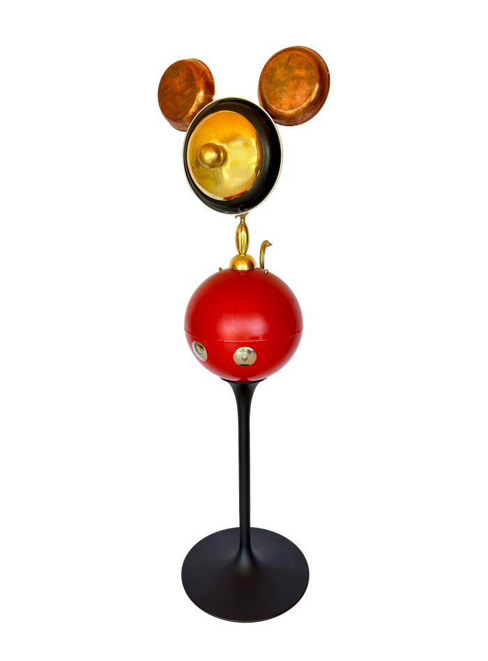 escultura on la cara daurada formada per una paella i les orelles son platets de balança, el cos es una bola bermeja molt gran i les cames una pues de talla de color negre